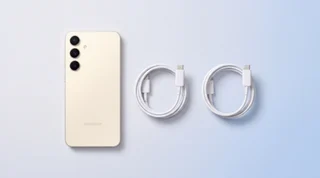 A black Samsung Galaxy S25 lying face down on a pale-grey surface with a white USB-C cable unplugged just in front of its charging port