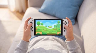 A white Nintendo Switch OLED lying flat with its screen off and a USB-C charging cable plugged into the port on its bottom edge, a soft blue glow at the connection