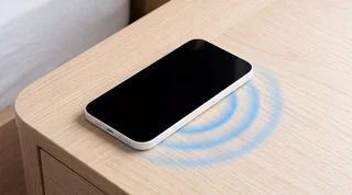 White iPhone 16 on a bedside table, screen still dark after a tap, faint blue rings on the wood beside it